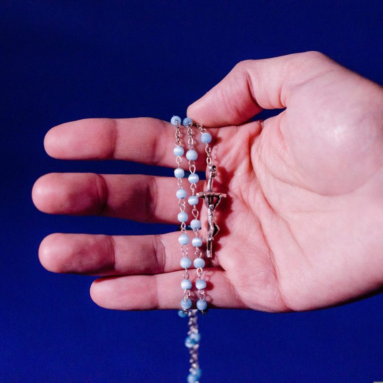 Eine Hand, die einen Rosenkranz hält. Das Kreuz zeigt Christus am Kreuz als Figur. Dahinter dunkelblauer Himmel.