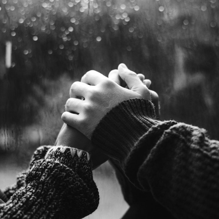 Zwei Hände, , die andere zwei Hände umschließen - dahinter eine Scheibe Glas, an der Regen herunterperlt.