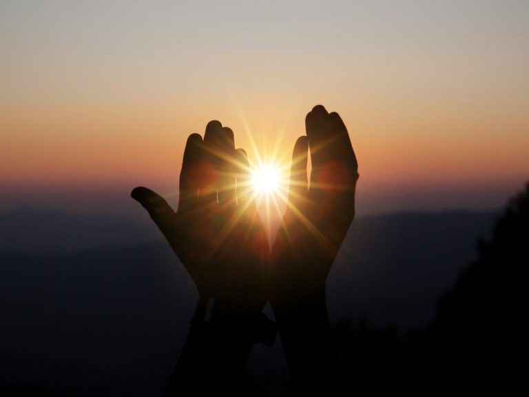 Zwei Hände die wie im Gebet nach oben gestreckt sind - dazwischen schaut die Sonne heraus  mit einem leuchtenden Strahlen. 