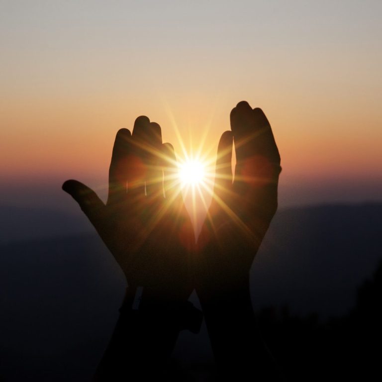 Zwei Hände - zum Himmel erhoben bei einem Sonnenaufgang oder Untergang. Zwischen den erhobenen Händen leuchtet die Sonne strahlend hindurch.
